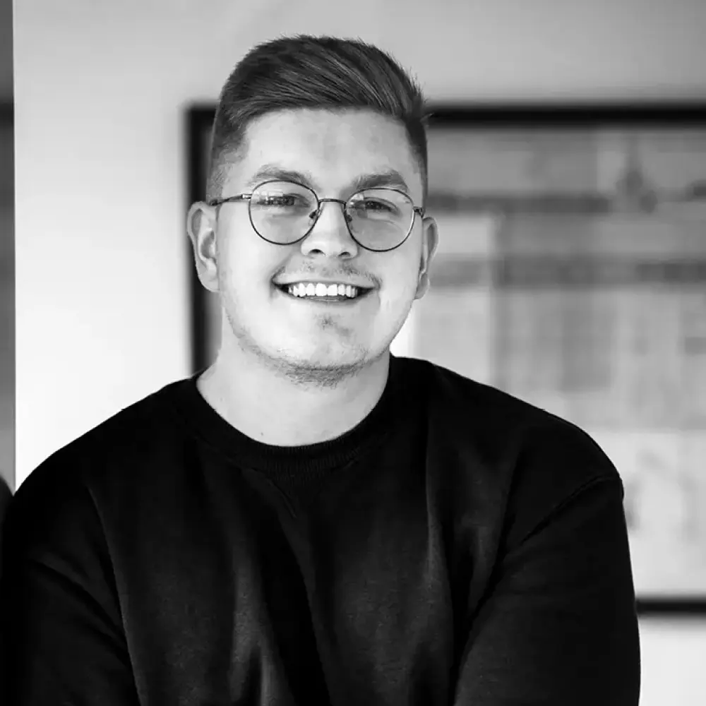 Junger Bauzeichner mit Brille und kurzem Haar, freundlich lächelnd vor Architekturplänen – STUDIO S Bocholt