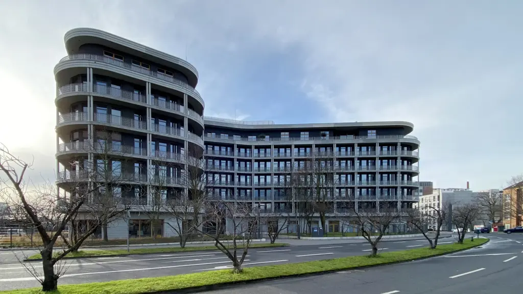 Panoramaaufnahme eines modernen Wohn- und Geschäftshauses mit geschwungener Fassade und umlaufenden Balkonen, aufgenommen vom Straßenraum.